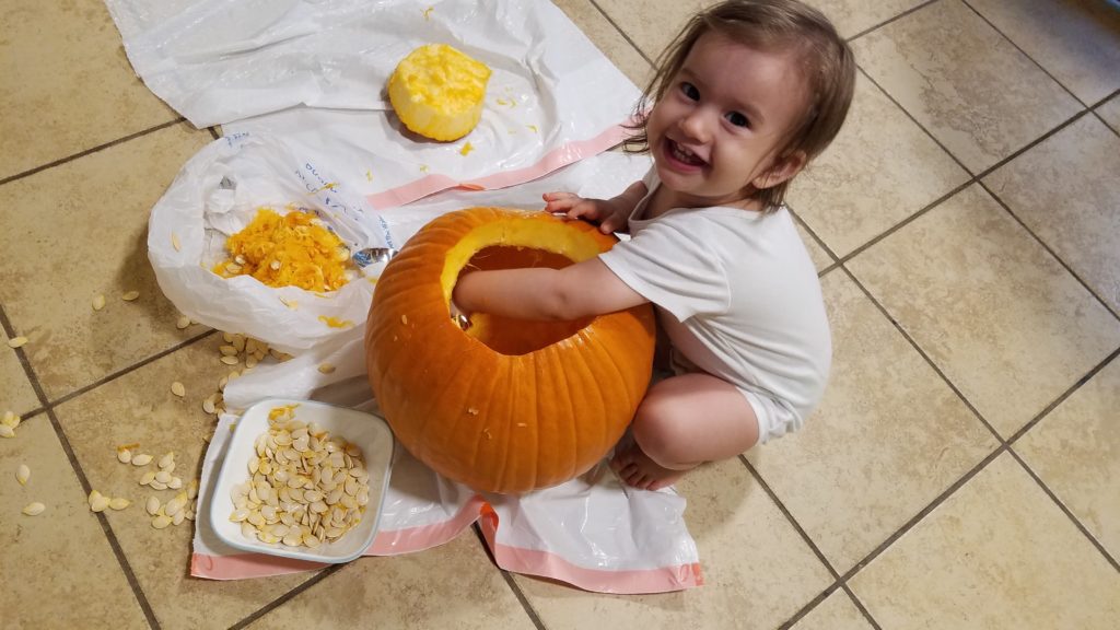 Mama Rissa's smiling toddler scooping guts out of her pumpkin exemplifying how to carve a pumpkin with your toddler.