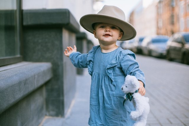 Toddler girl in a dress wearing a hat and holding a teddy bear.